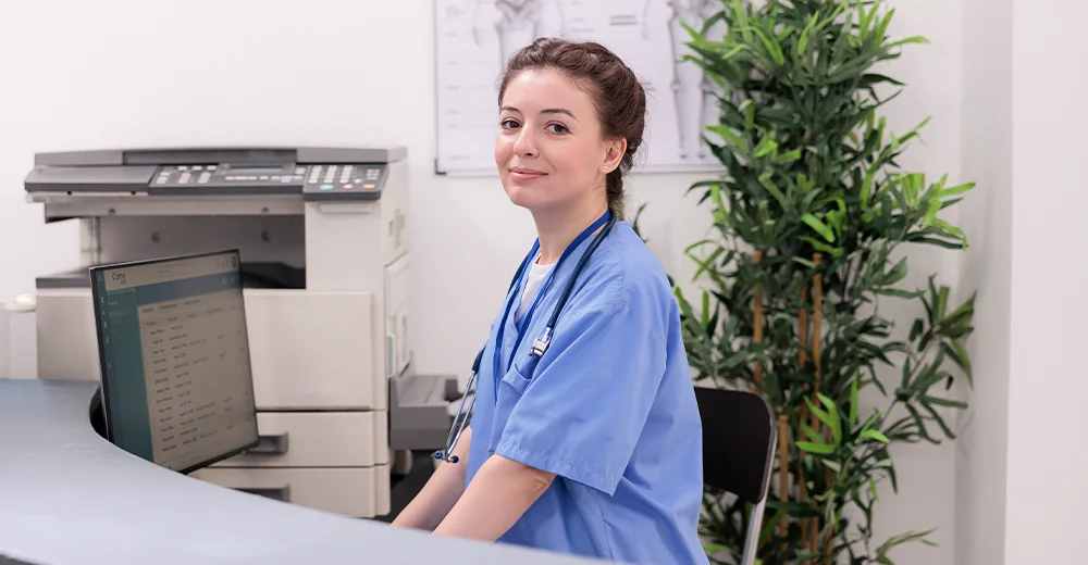 A medical administrative assistant seated at the reception desk of a medical office.