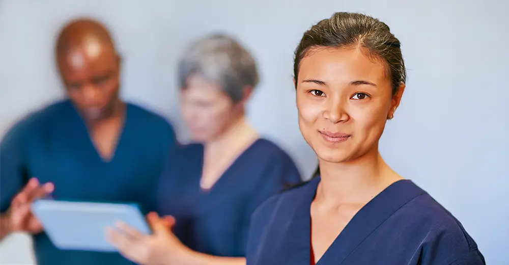 Hospital unit clerk in the foreground of a medical setting, with two team members reviewing a file in the background.